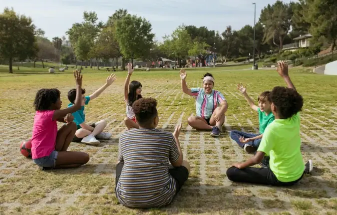 Children sitting in a circle on the grass, raising their hands with a coach
